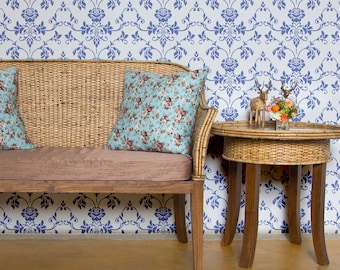 Könnte beinhalten: Ein Korbsofa mit zwei floral gemusterten Kissen steht vor einer blau-weißen Tapeten mit Damastmuster. Rechts vom Sofa steht ein kleiner Korb-Beistelltisch mit Glasplatte. Auf dem Tisch stehen eine kleine Hirschfigur und eine Vase mit Blumen.