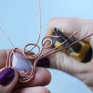 May include: A close-up of a copper wire wrapped around a light purple and orange agate stone. The wire is being shaped into a decorative design with pliers.