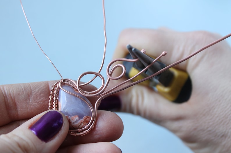 May include: A close-up of a copper wire wrapped around a light purple and orange agate stone. The wire is being shaped into a decorative design with pliers.
