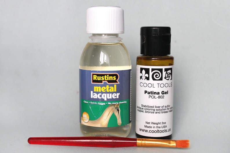 May include: Two bottles of metal finishing products. A clear bottle of Rustins metal lacquer and a brown bottle of Cool Tools Patina Gel. Both bottles are labeled with product information. A red and gold paintbrush is in the foreground.