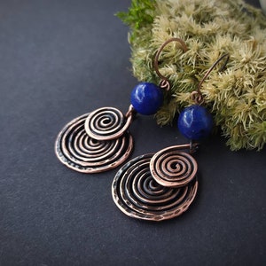 以下が含まれることがあります： 螺旋模様と濃い青色の宝石が付いた銅製のイヤリングのペア。イヤリングは暗い背景にぶら下がっています。