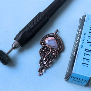 May include: A copper wire wrapped pendant with a light blue and brown agate stone. The pendant is on a blue surface with a black rotary tool and a blue box labeled "Dialux Bleu" with the text "Bleu de Paris".