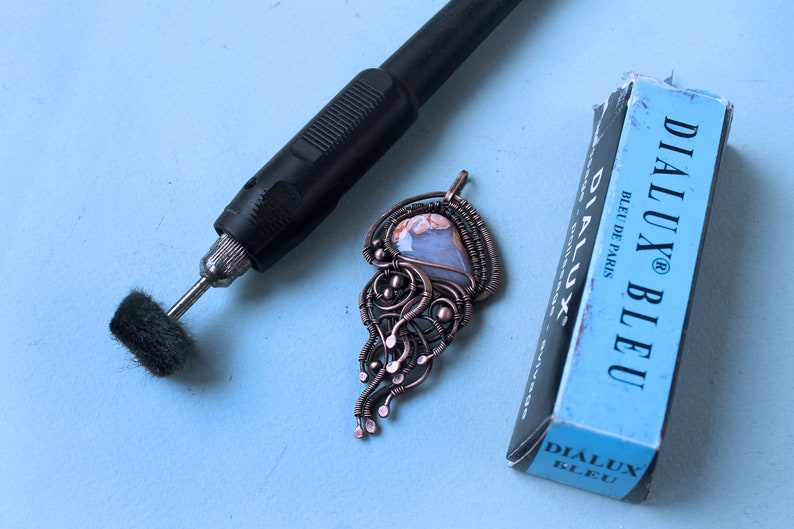 May include: A copper wire wrapped pendant with a light blue and brown agate stone. The pendant is on a blue surface with a black rotary tool and a blue box labeled "Dialux Bleu" with the text "Bleu de Paris".