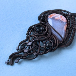 May include: A brown wire wrapped pendant with a light gray and pink agate stone. The wire is twisted and formed into a delicate design with black beads.