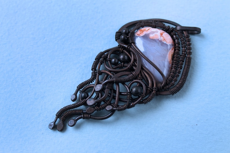 May include: A brown wire wrapped pendant with a light gray and pink agate stone. The wire is twisted and formed into a delicate design with black beads.