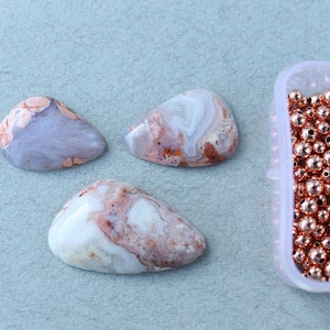 May include: Three white and pink agate cabochons with brown and orange swirls, next to a container of copper-colored beads.