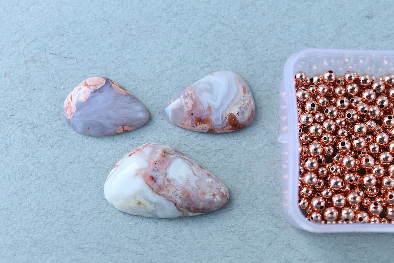 May include: Three white and pink agate cabochons with brown and orange swirls, next to a container of copper-colored beads.