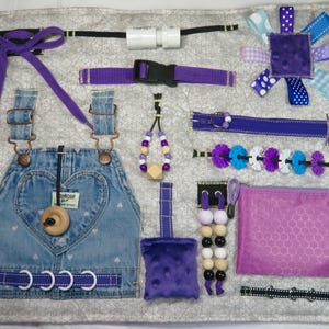 May include: A sensory activity board with various textures and fasteners. Features denim overalls with a heart design, purple ribbons, buckles, zippers, beads, and a small pouch. The board is designed to stimulate tactile exploration and fine motor skills.