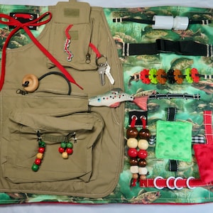 May include: A sensory activity board featuring a tan vest with pockets, red accents, and various tactile elements. Includes keys, wooden beads, a toy fish, and a green and red patterned background. Designed for sensory exploration and fine motor skill development.