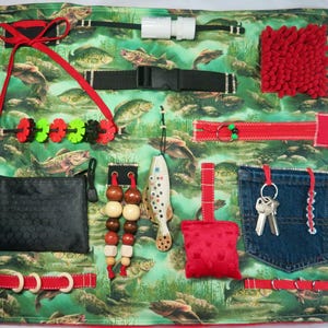 May include: Sensory activity board with various textures and interactive elements. Features a green fabric with a fish pattern, a red zipper, a denim pocket with keys, and a black buckle. Includes a red ribbon, a black wallet, and wooden beads.