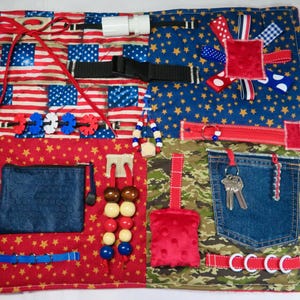 May include: A sensory activity mat with various textures and interactive elements. Features include a zipper, buckle, keys, beads, and a small pocket. The mat incorporates patriotic patterns with red, white, and blue colors.