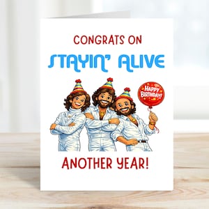 Puede incluir: Tarjeta de felicitación de cumpleaños con el texto "CONGRATS ON STAYIN' ALIVE ANOTHER YEAR!" en rojo y azul. La tarjeta presenta una ilustración de dibujos animados de tres hombres con trajes blancos y sombreros de fiesta, uno sosteniendo un globo de "Happy Birthday!"