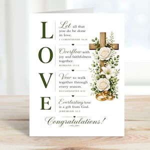 Könnte beinhalten: Eine weiße Hochzeitskarte mit dem Wort "LOVE" in großen grünen Buchstaben. Die Karte zeigt ein Holzkreuz, das mit weißen Rosen, Lilien und goldenen Eheringen geschmückt ist. Der Text enthält Bibelverse und "Glückwunsch!"