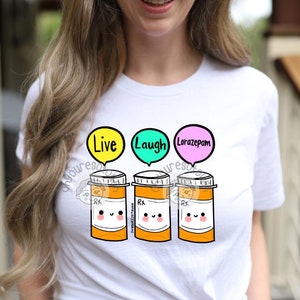 May include: White t-shirt with a graphic of three orange prescription pill bottles with smiling faces. Above each bottle are speech bubbles with the words "Live", "Laugh", and "Lorazepam". The shirt is worn by a person.