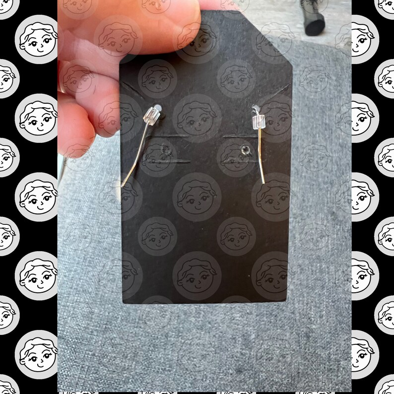 May include: A pair of silver earrings with clear crystal accents. The earrings are displayed on a black card with a repeating pattern of cartoon faces.