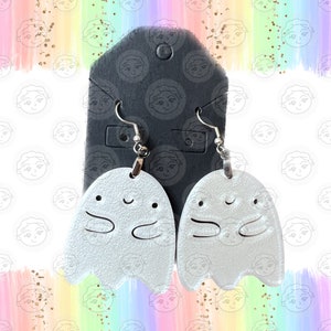 May include: A pair of silver ghost earrings with a smiling face and outstretched arms. The earrings are hanging from silver hooks.
