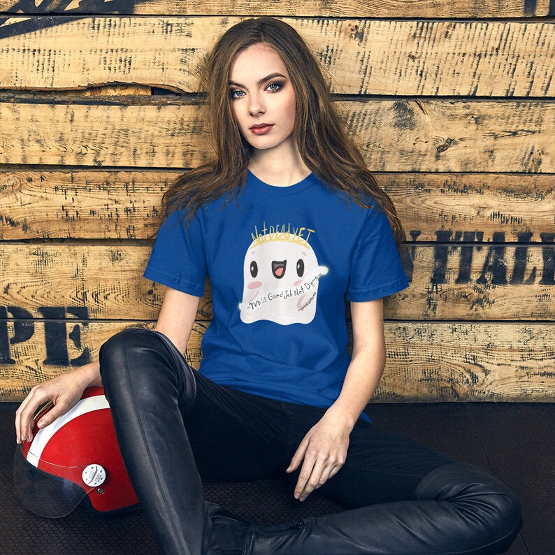 May include: A woman wearing a royal blue t-shirt with a white ghost graphic. The ghost has a gold crown and the text "Material Vet" above it. The ghost is smiling and has the text "Y'all is Good, I'll Not Die" written on it.