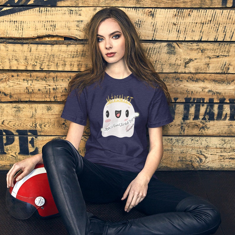 May include: A woman wearing a navy blue t-shirt with a white ghost graphic. The ghost has a gold crown and the text "Not Real Vet" above it. The ghost is smiling and has the text "Y'all is Good, I'll Not Die" written on it.