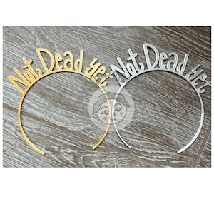 May include: Two headbands with the text "Not Dead Yet" in gold and silver. The headbands are on a wooden surface.