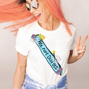 May include: White t-shirt with a blue sash that says "Mx. Not Dead Yet" with a crown, flowers, and a wand.