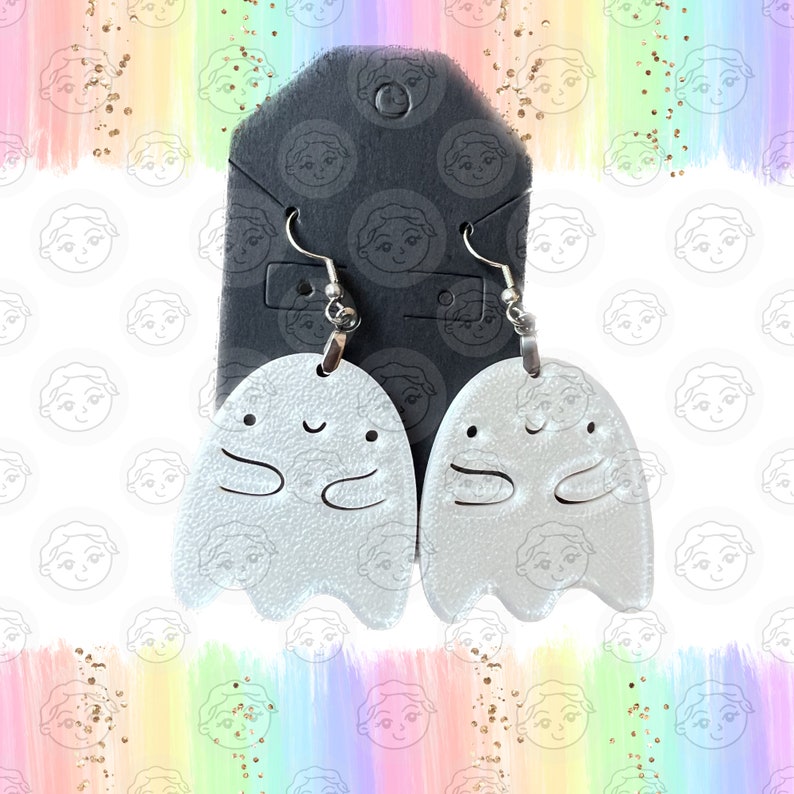 May include: A pair of silver ghost earrings with a smiling face and arms. The earrings are hanging from a black card.