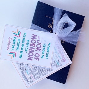 May include: A white bookmark with a colorful design and the text "PROMISE THAT YOU READ THE BOOK OF MORMON EVERY DAY YOU WILL MAKE BETTER DECISIONS EVERY DAY." - Russell M. Nelson. The bookmark is tied to a blue book with a white ribbon.