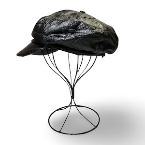 May include: A black leather newsboy cap featuring a textured, crinkled surface finish and a short, stiff brim. The cap is constructed from multiple stitched panels that meet at the top center, creating a rounded, voluminous shape.