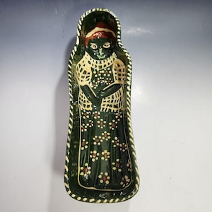 May include: A ceramic serving dish in the shape of a woman. The dish is dark green with a floral pattern and a cream-colored lattice design. The woman's face is black with red hair and a green smile. The dish is approximately 12 inches long.