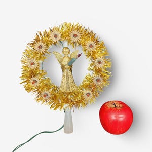 May include: A gold tinsel wreath tree topper featuring a central angel figure with wings and a dress made of gold mesh. The wreath is decorated with small star-shaped light covers and a single red apple is positioned to the right of the topper.