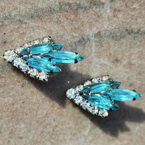 May include: A pair of vintage earrings featuring a cluster of light blue, teardrop-shaped crystals. The crystals are surrounded by smaller, clear, round crystals set in a silver-toned metal. The earrings have a classic, elegant design.