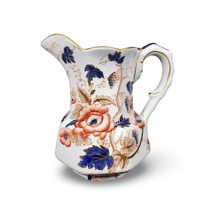 May include: A white ceramic pitcher featuring a decorative pattern of large orange flowers, dark blue leaves, and brown vines. The pitcher has a curved handle, a scalloped rim, and gold-colored trim along the edges of the handle and the top rim.