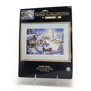 May include: A Dimensions The Gold Collection counted cross stitch kit titled Sleigh Ride by Nicky Boehme. The packaging features a winter village scene with a horse-drawn sleigh, snow-covered houses, and a church. It measures 15 inches by 11 inches.