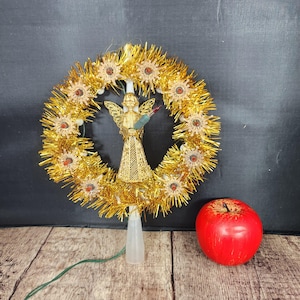 May include: A vintage Christmas tree topper featuring a gold angel figure holding a small object, centered within a gold tinsel wreath. The wreath is adorned with small, decorative elements. A red apple sits nearby. The topper is on a clear plastic base.