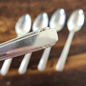 Könnte beinhalten: Ein Set aus vier silberfarbenen Metalllöffeln mit polierter Oberfläche. Der Griff des vorderen Löffels zeigt ein rechteckiges Ende mit dezenten geprägten Linien- und Punktverzierungen an den Kanten, während die anderen drei Löffel unscharf sind.