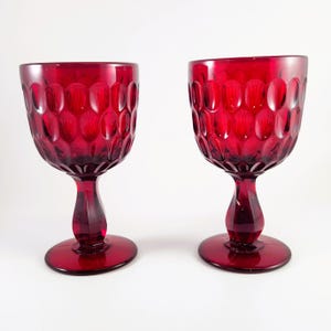 May include: Two matching deep red glass goblets featuring a prominent honeycomb or thumbprint pattern on the bowl. Each glass has a thick, faceted stem and a wide, circular base, crafted from translucent ruby-colored glass with a smooth finish.