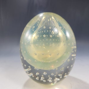 May include: A clear, egg-shaped glass paperweight with a yellow and blue interior design. The glass is filled with small bubbles, creating a unique visual texture. The paperweight is a decorative item.