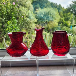 May include: Three ruby red glass pitchers with clear glass handles. The pitchers have a textured surface and varying shapes, with one having a flared rim. They are displayed on a clear acrylic shelf in front of a window.