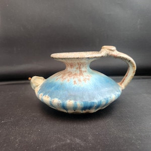 May include: A ceramic oil lamp with a handle and spout. The lamp features a blue and brown glaze, with a light blue body and a brown rim. The handle and spout are a light brown color. The lamp is likely handmade and has a unique, rustic appearance.