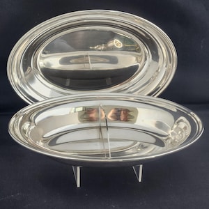 May include: Two silver serving dishes. The top dish is a large oval platter. The bottom dish is a smaller oval serving bowl with three small feet. Both dishes have a shiny, reflective surface.