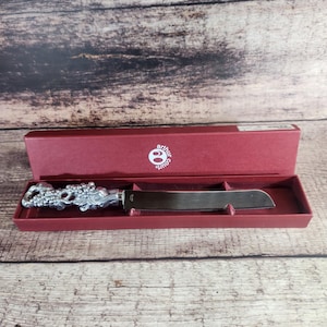 May include: A silver-toned metal knife with a decorative handle featuring a sculpted grape and vine motif, presented in a rectangular maroon box with the Arthur Court logo printed in white on the interior lid of the packaging.