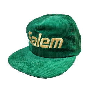 May include: A forest green corduroy baseball cap featuring the word Salem embroidered in gold thread across the front panel. The hat has a structured crown and a flat brim, both made from the same textured corduroy fabric with a consistent finish.
