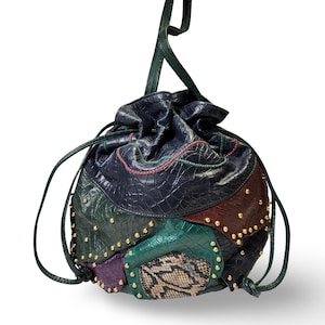 May include: A drawstring bucket bag featuring a patchwork design of dark blue, green, purple, and brown textured panels with a snakeskin pattern section. The bag is accented with small gold-tone metal studs along the seams and has a long shoulder strap.