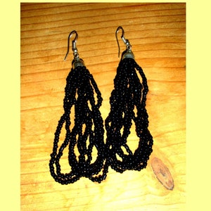 May include: A pair of black beaded earrings with silver-colored hooks. The earrings feature multiple strands of small, round beads, creating a cascading effect. The beads are a deep black color, and the earrings are displayed against a wooden background.