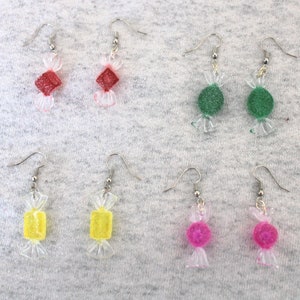 May include: Six pairs of candy-shaped earrings. The earrings are made of clear plastic with a candy-shaped charm. The charms are red, green, yellow, and pink.