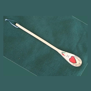 Puede incluir: Una cuchara de madera con un diseño pintado. La cuchara presenta un corazón rojo, un chile verde y las palabras "I love chili stew". La cuchara tiene un pequeño lazo en el extremo del mango.