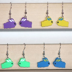 May include: Four pairs of dangle earrings featuring a bunny rabbit design. The earrings are available in purple, yellow, green, and blue.