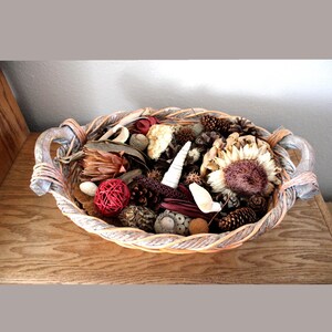 May include: An oval woven basket with two handles containing a decorative assortment of dried natural elements including pinecones, a large dried sunflower, a red rattan sphere, various seed pods, and small white shells arranged inside.