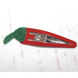 Op de afbeelding: Een rode haarclip in de vorm van een chilipeper, met een groene vilten steel. De clip is gemaakt van metaal en heeft een eenvoudig ontwerp. Dit accessoire is geschikt voor diverse kapsels.