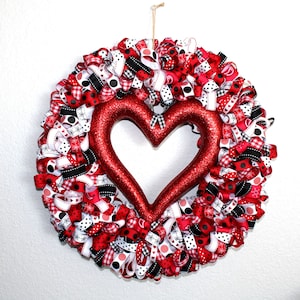 May include: A Valentine's Day wreath with a large, glittery red heart in the center. The wreath is made of red, white, and black ribbons with various patterns, including polka dots, gingham, and hearts. It hangs against a white wall.
