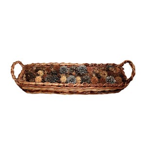 May include: A rectangular woven brown wicker tray with two curved handles on the ends. The tray is filled with a collection of small, dark brown pinecones and light tan spiky seed pods, creating a textured, natural arrangement within the basket.